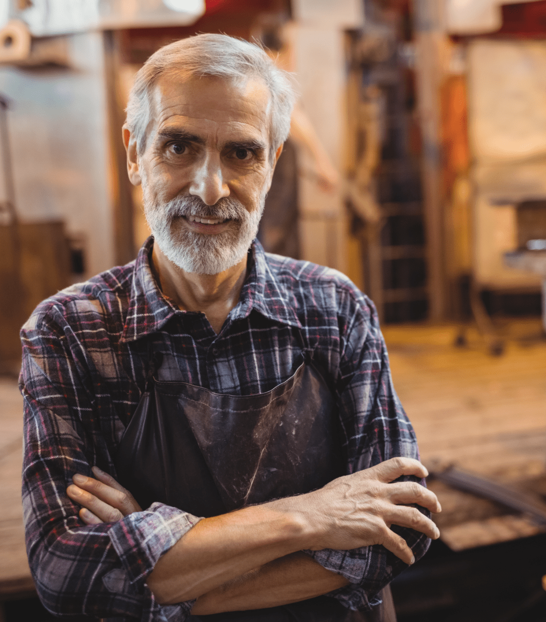 Ein älterer Mann mit weißen Haar und Bartwuchs trägt ein kariertes Hemd, sowie eine graue Latzhose und lächelt in der Handwerkerwerkstatt mit verschränkten Armen in die Fotokamera