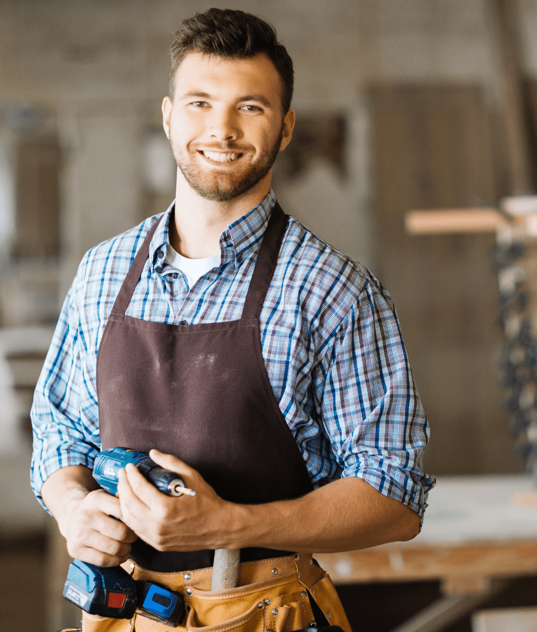 Attraktiver junger Mann lächelt mit kariertem Arbeitshemd, brauner Schürze und Werkzeuggürtel in die Fotokamera, während er eine Bohrmaschine in der Hand hält